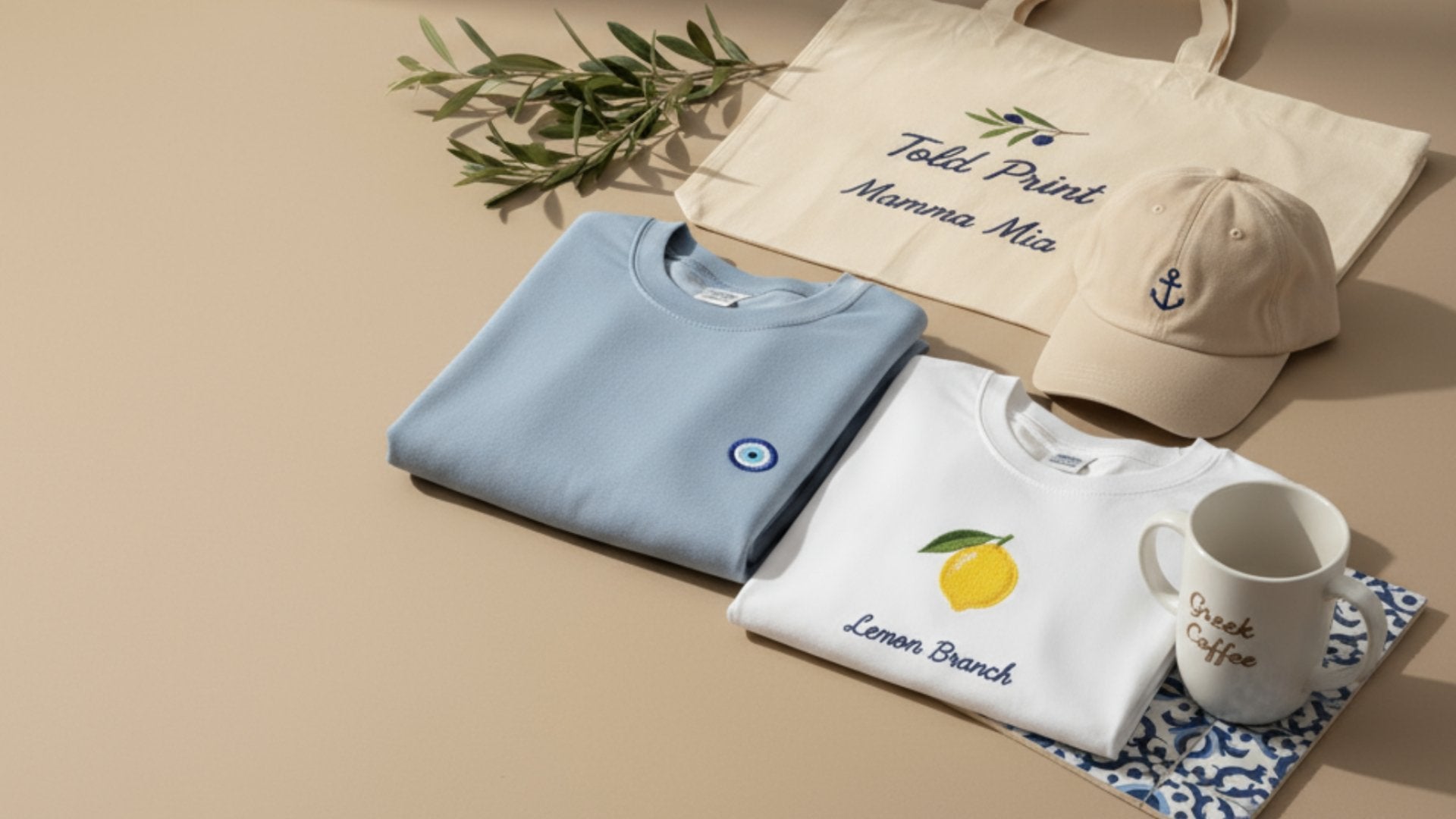 Flat lay of ToldPrint Mediterranean items — embroidered sweatshirts, tote, cap, and mug with olive branch styling.