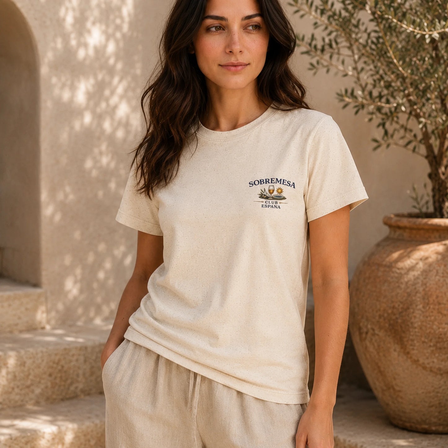 A woman wearing a cream-colored short-sleeve t-shirt with a small 'SOBREMESA' graphic on the left chest, paired with matching cream pants, standing in a sunlit rustic setting with a large terracotta pot.
