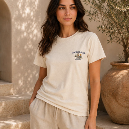 A woman wearing a cream-colored short-sleeve t-shirt with a small 'SOBREMESA' graphic on the left chest, paired with matching cream pants, standing in a sunlit rustic setting with a large terracotta pot.