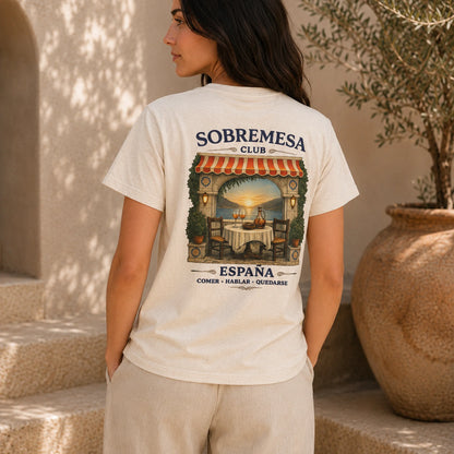 A woman wearing a cream-colored t-shirt with a large graphic print on the back, standing in a sunlit outdoor setting with stone steps and a potted plant.