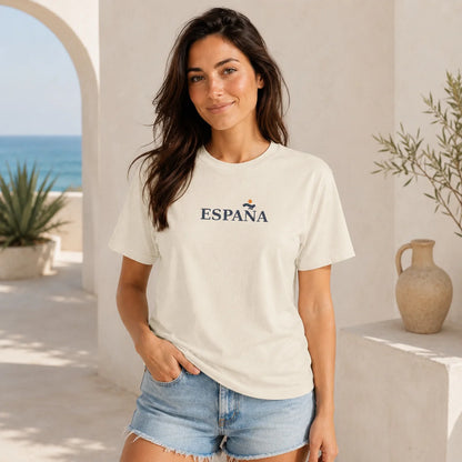 A woman wearing a cream-colored t-shirt with 'ESPAÑA' printed in navy blue across the chest, paired with light blue denim shorts, standing in a sunny, Mediterranean-style outdoor setting with a white stucco wall, archway, and sea in the background.