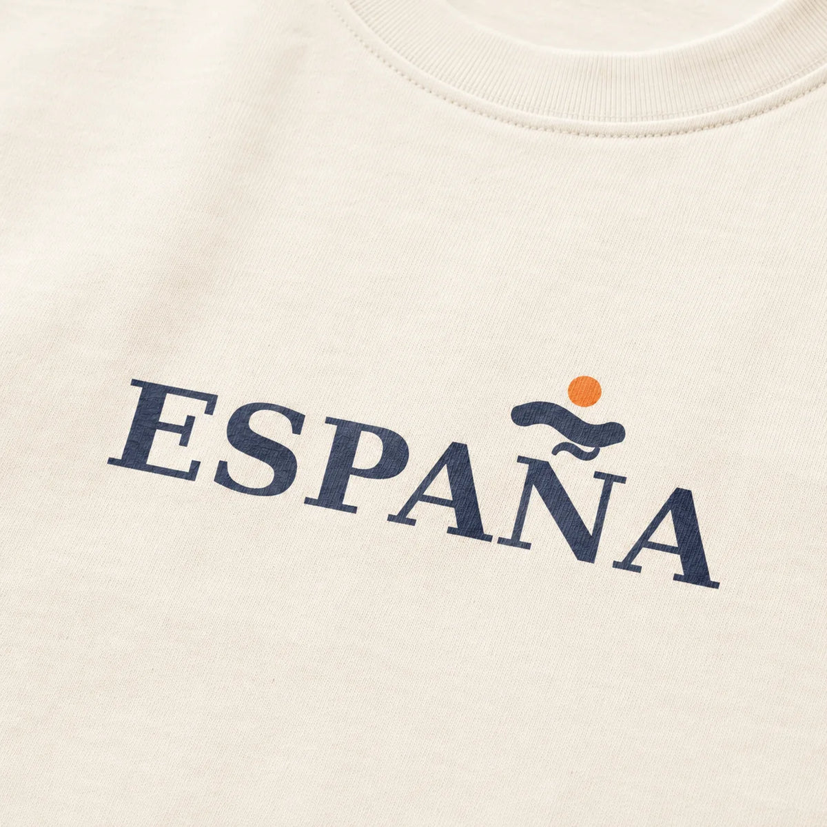 Close-up of a cream-colored t-shirt featuring the word 'ESPAÑA' printed in navy blue across the chest, with a small orange sun and blue wave graphic above the 'A'.