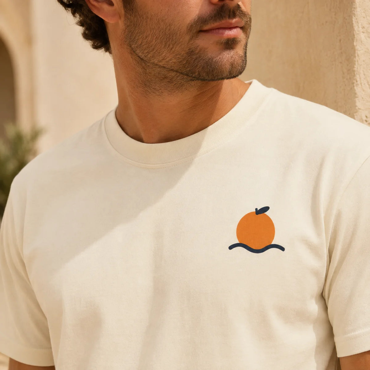 A man wearing a cream-colored crewneck t-shirt with a small graphic of an orange on the left chest, set against a warm beige background.