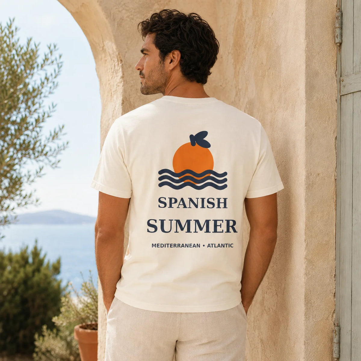 A man stands outdoors in front of a beige stone archway wearing a cream-colored t-shirt with a graphic on the back. The graphic features an orange sun, blue waves, and the text 'SPANISH SUMMER' with 'MEDITERRANEAN • ATLANTIC' underneath.