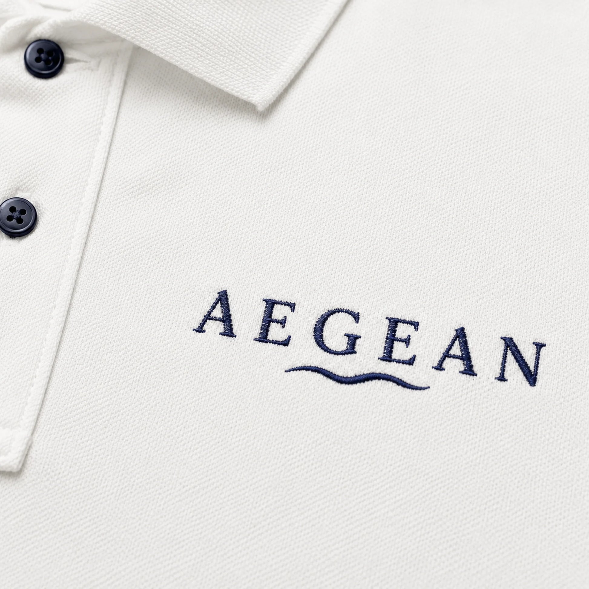 Close-up of a white polo shirt with the brand name 'AEGEAN' embroidered in navy blue on the chest, featuring a two-button placket.