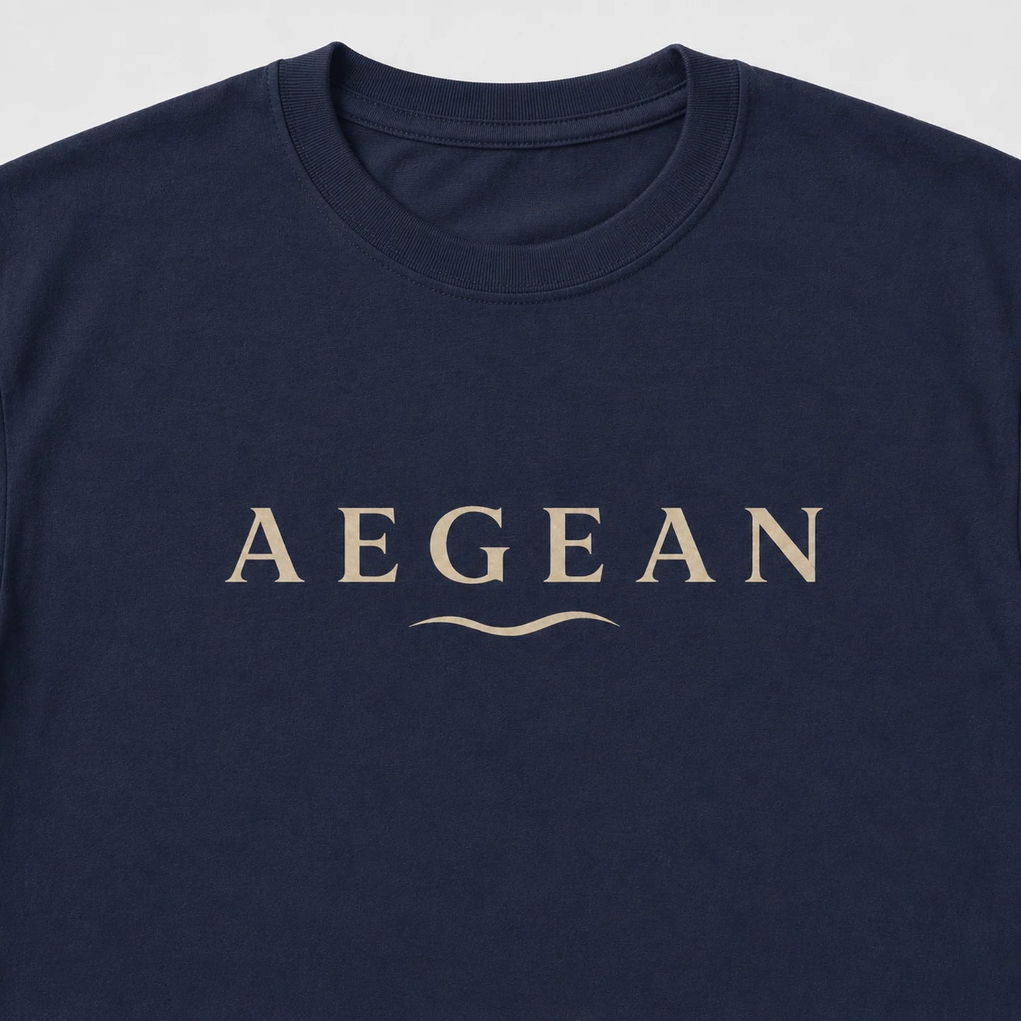 A close-up of a navy blue crewneck t-shirt featuring the word 'AEGEAN' printed in a beige serif font across the chest, with a small wavy line underneath.
