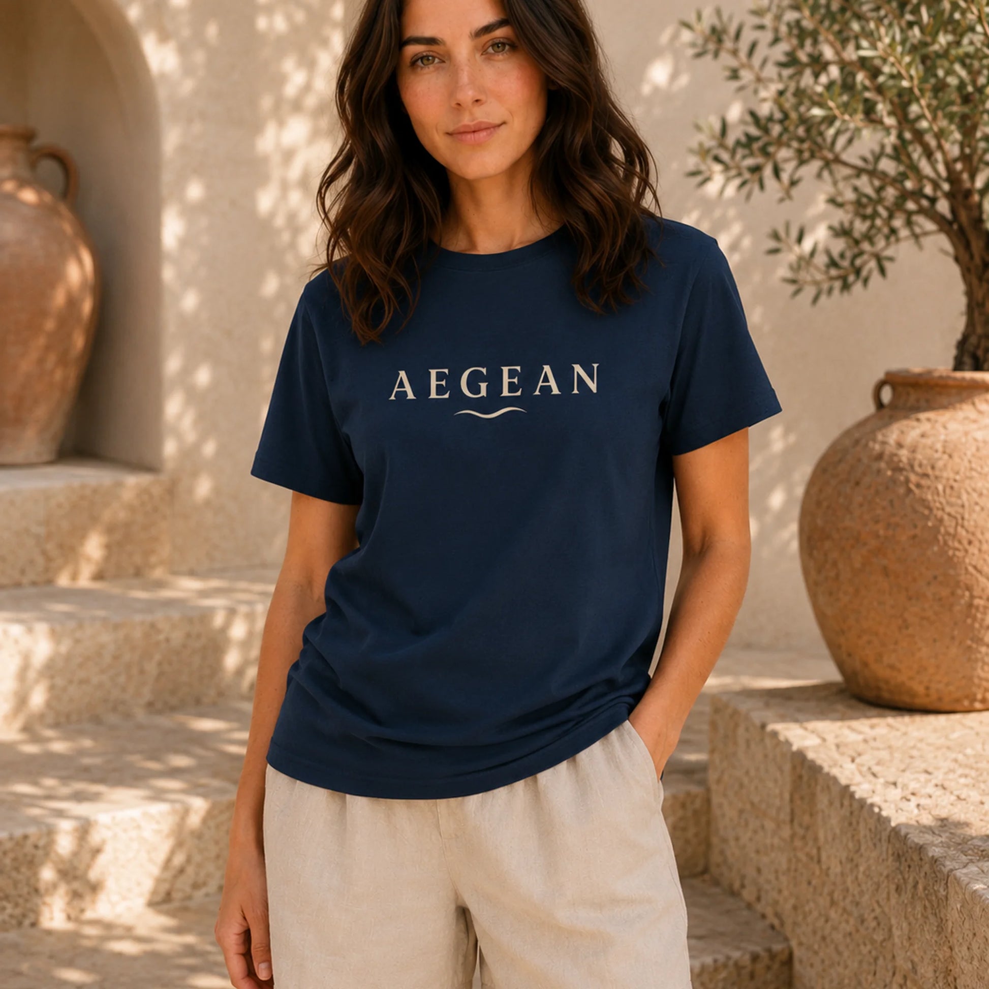 A woman wearing a navy blue crewneck t-shirt with 'AEGEAN' printed in white across the chest, paired with light beige shorts, standing in a sunlit outdoor setting with stone steps and large clay pots.