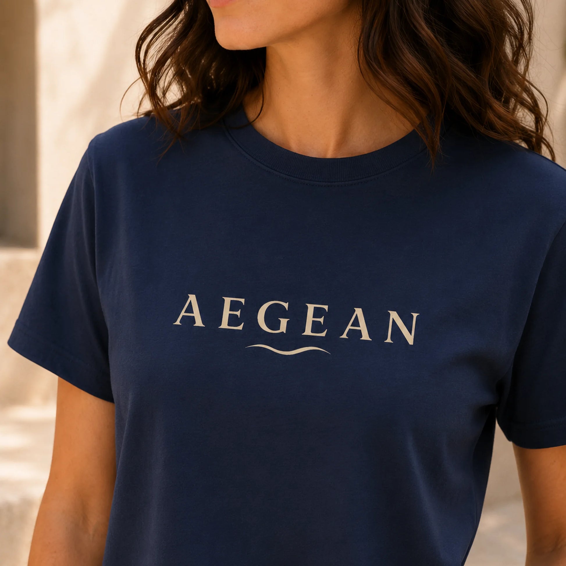 TP HERO: A woman wearing a navy blue crewneck t-shirt with the word 'AEGEAN' printed in beige across the chest, shown in a close-up shot.
