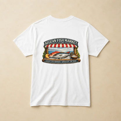 A white short-sleeve crewneck t-shirt laid flat on a light beige background, featuring a colorful graphic print on the front with a fish market scene and the text 'AEGEAN FISH MARKET CLUB'.