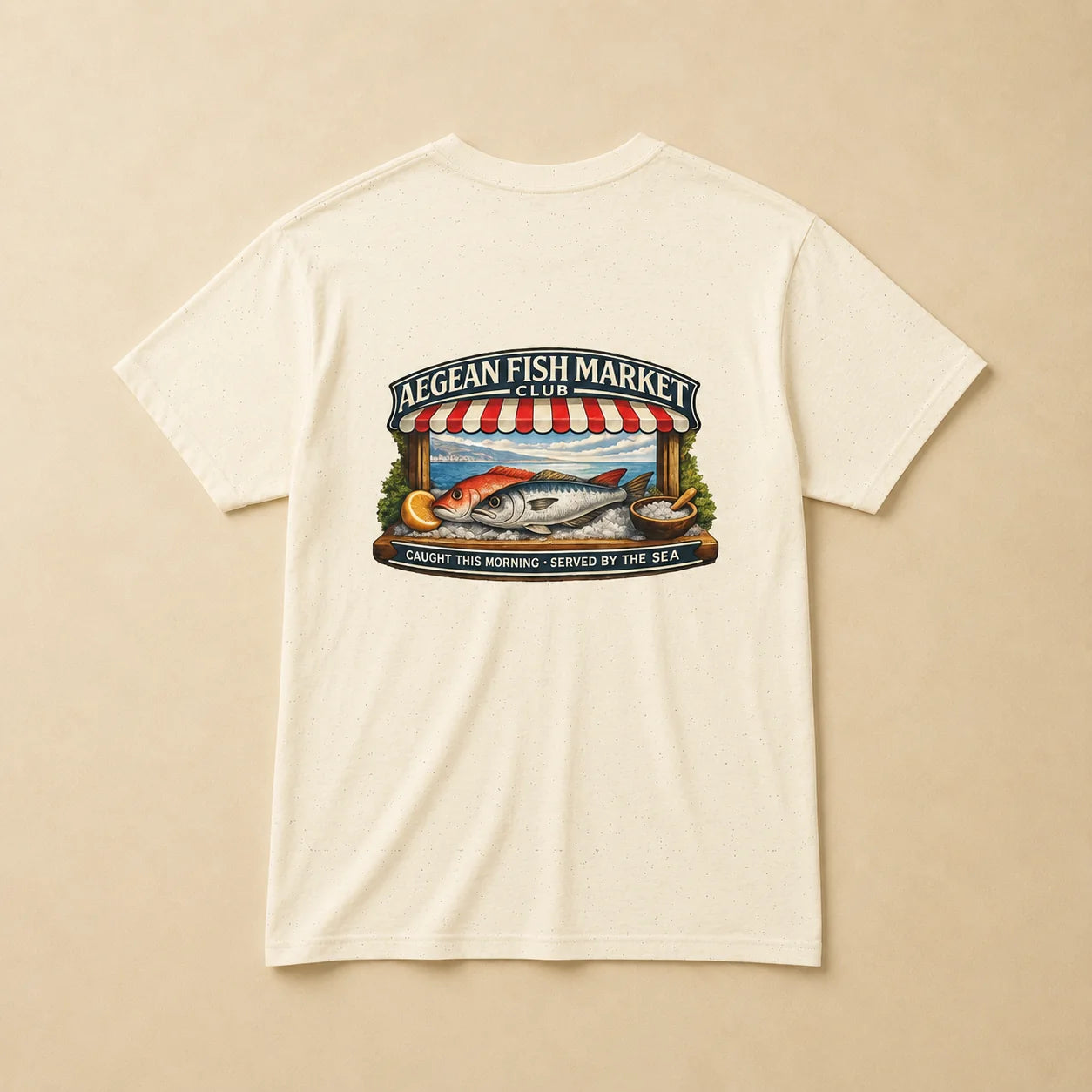 An off-white short-sleeve t-shirt with a colorful graphic print on the front depicting a fish market scene with a striped awning, fish, and text reading 'AEGEAN FISH MARKET' and 'CAUGHT THIS MORNING - SERVED BY THE SEA'.