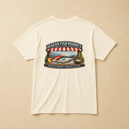 An off-white short-sleeve t-shirt with a colorful graphic print on the front depicting a fish market scene with a striped awning, fish, and text reading 'AEGEAN FISH MARKET' and 'CAUGHT THIS MORNING - SERVED BY THE SEA'.