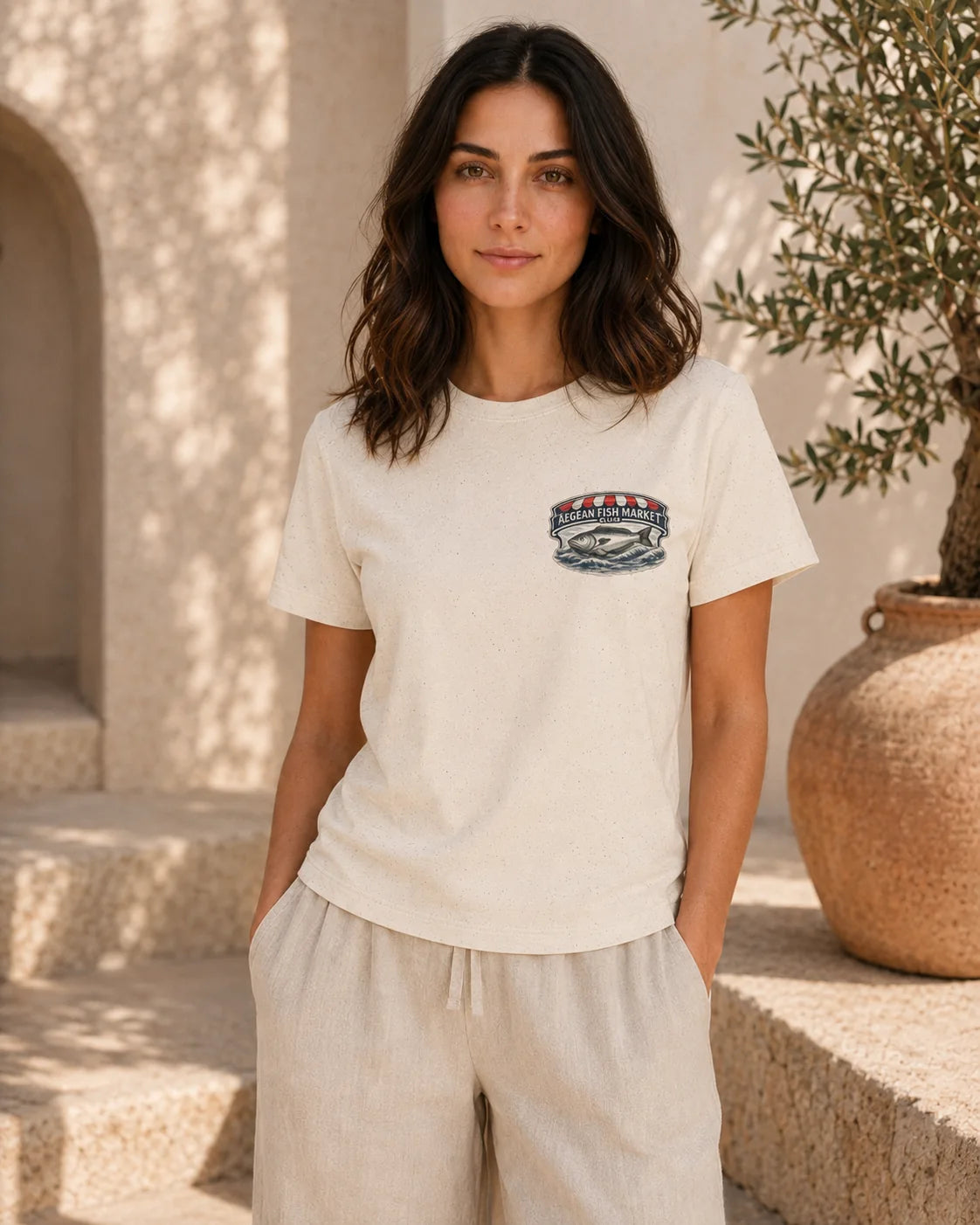 A woman wearing a cream-colored short-sleeve t-shirt with a small embroidered logo on the left chest, paired with matching beige linen pants, standing in a sunlit outdoor setting with stone steps and a potted olive tree.