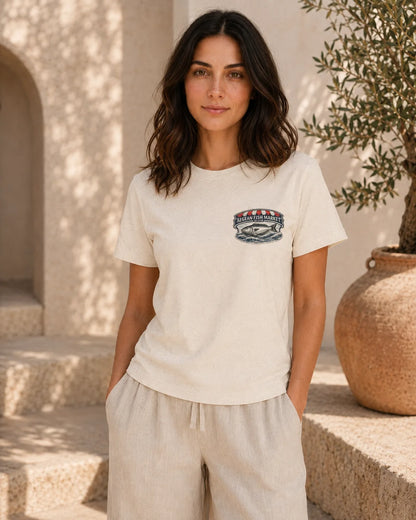 A woman wearing a cream-colored short-sleeve t-shirt with a small embroidered logo on the left chest, paired with matching beige linen pants, standing in a sunlit outdoor setting with stone steps and a potted olive tree.