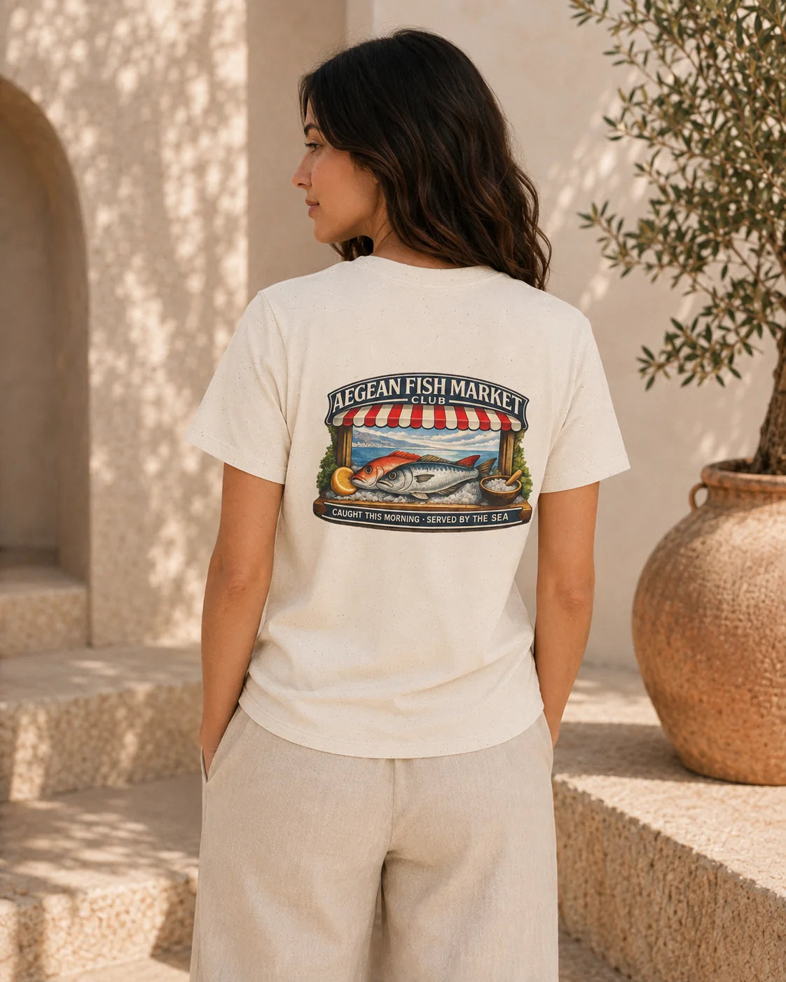 A woman wearing a cream-colored t-shirt with a colorful graphic of a fish market on the back, standing outdoors in a sunlit Mediterranean-style setting.