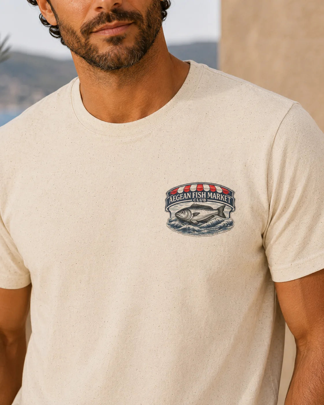 A man wearing a light beige crewneck t-shirt with a 'Mega Fish Market' fish graphic printed on the left chest, set against a blurred outdoor background.