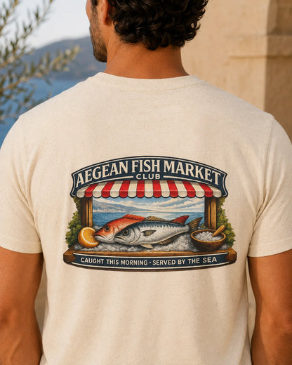 TP HERO: A man viewed from behind wearing a cream-colored t-shirt with a large graphic print on the back. The graphic features a fish market scene with the text 'AEGEAN FISH MARKET CLUB' and 'CAUGHT THIS MORNING - SERVED BY THE SEA'.