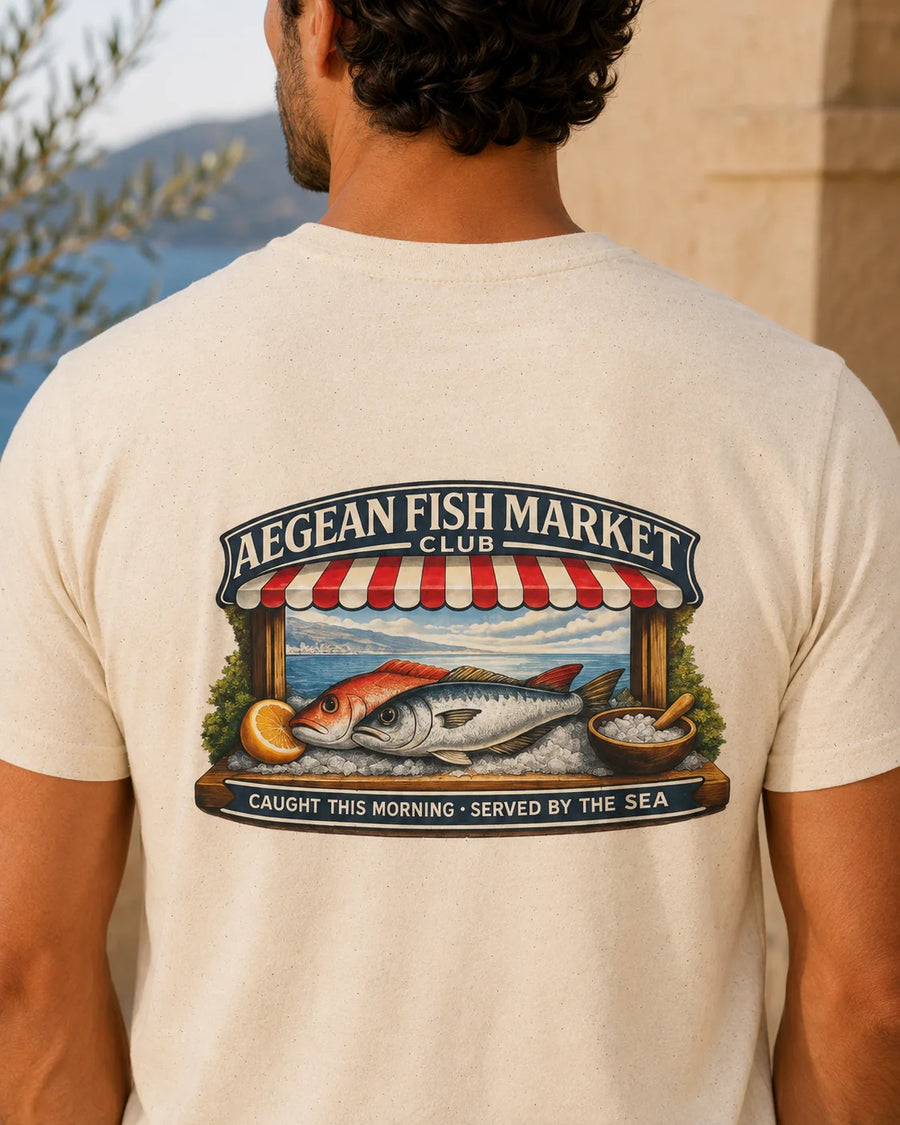 TP HERO: A man viewed from behind wearing a cream-colored t-shirt with a large graphic print on the back. The graphic features a fish market scene with the text 'AEGEAN FISH MARKET CLUB' and 'CAUGHT THIS MORNING - SERVED BY THE SEA'.