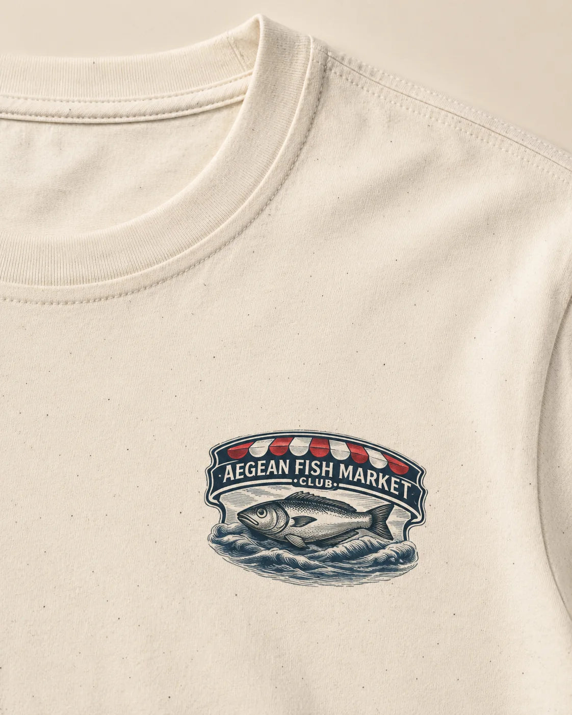 Close-up of an off-white crewneck t-shirt featuring a vintage-style graphic on the chest that reads 'AEGEAN FISH MARKET CLUB' with a fish illustration.