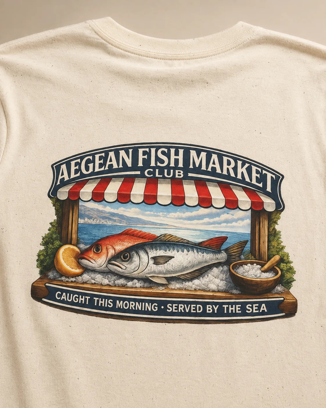Close-up of a cream-colored t-shirt featuring a large graphic print of a fish market scene with a red and white striped awning, a large fish, and the text 'AEGEAN FISH MARKET CLUB' and 'CAUGHT THIS MORNING - SERVED BY THE SEA'.