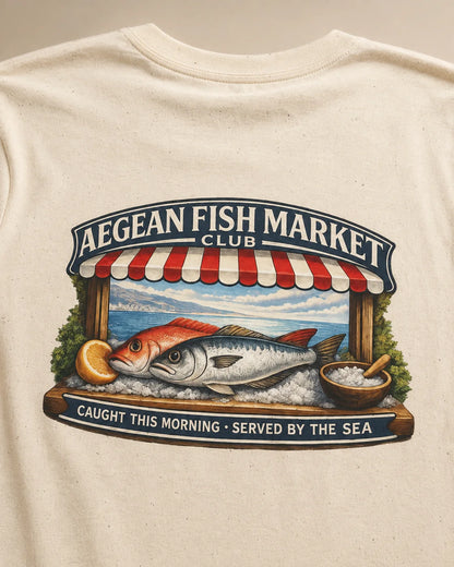 Close-up of a cream-colored t-shirt featuring a large graphic print of a fish market scene with a red and white striped awning, a large fish, and the text 'AEGEAN FISH MARKET CLUB' and 'CAUGHT THIS MORNING - SERVED BY THE SEA'.
