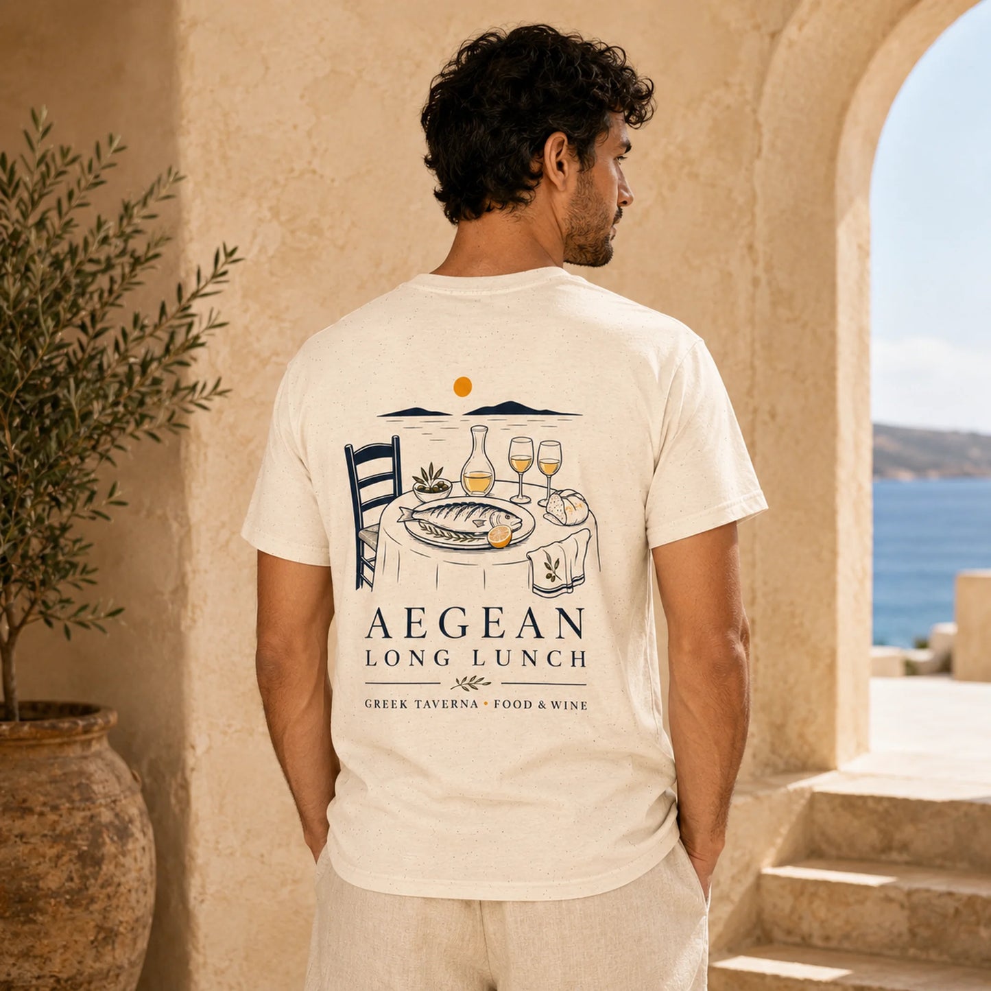 A man wearing a cream-colored t-shirt with a detailed graphic print on the back, standing in a Mediterranean-style setting with a stone archway and sea view in the background.