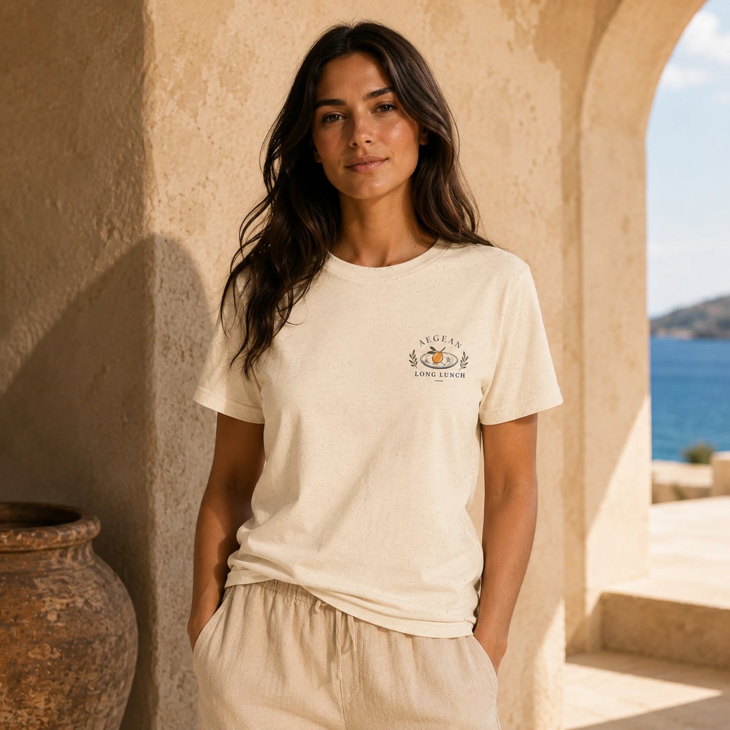 A woman stands in a sunlit stone corridor wearing a cream short-sleeve T-shirt with a small multicolor chest graphic and light beige drawstring pants. The background shows textured stucco walls, a large陶 pot, and an open arch leading to the sea.