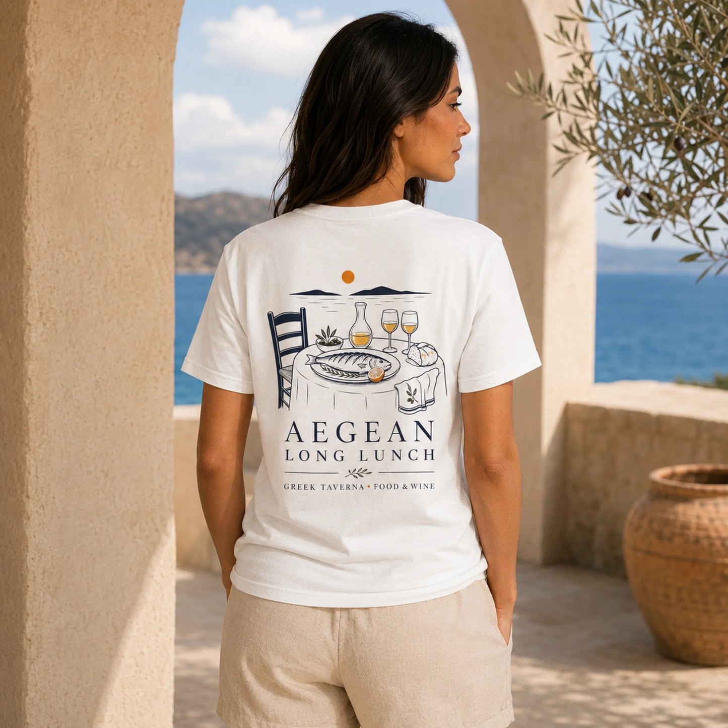 A woman stands outdoors in a beige stone archway, wearing a white graphic T-shirt with a dark blue and orange illustration of a table setting and the text 'AEGEAN LONG LUNCH'.