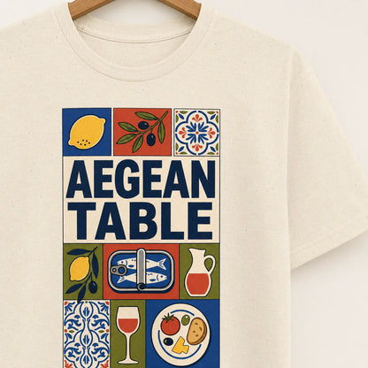 Beige t-shirt with 'Aegean Table' graphic design featuring lemons, olives, and other elements on a hanger.