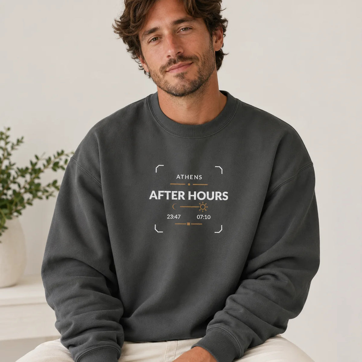 A man wearing a dark charcoal gray crewneck sweatshirt with a white and orange embroidered design that reads 'ATHENS AFTER HOURS 23:47 07:10'.