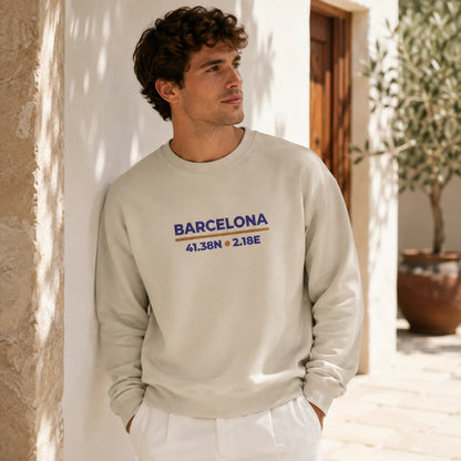 A man wearing a light beige crewneck sweatshirt with a blue and orange 'BARCELONA' graphic on the chest, standing outdoors against a white stucco wall.