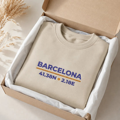 A beige crewneck sweatshirt with a blue and gold Barcelona embroidered design, neatly folded inside a brown cardboard gift box lined with white tissue paper.