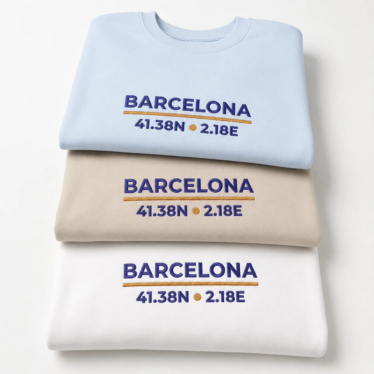 Three folded Barcelona Coordinates Light Edition crewneck sweatshirts in Light Blue, Sand and White, showing the navy and gold embroidered city-coordinate design.
