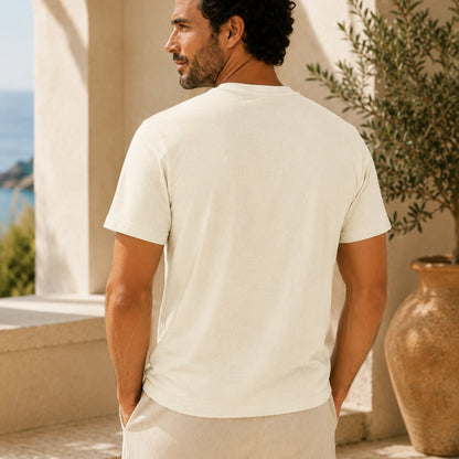 A man wearing a plain off-white short-sleeve t-shirt, shown from the back in a sunny outdoor setting with a large terracotta pot and a distant view of the sea.