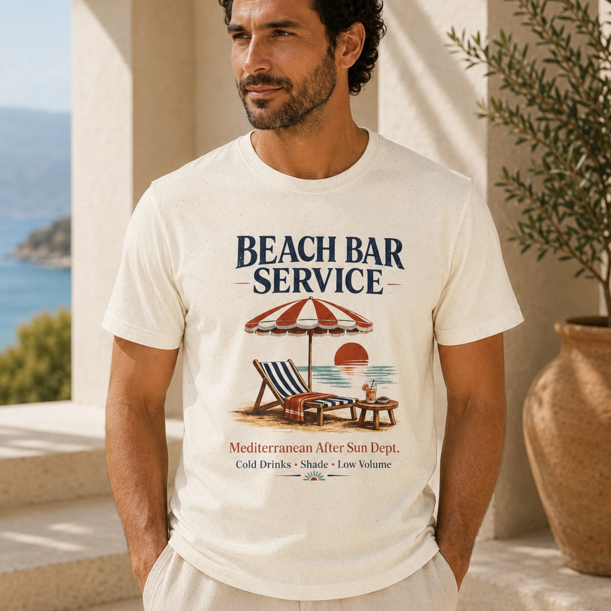 A man wearing a cream-colored graphic t-shirt with a 'Beach Bar Service' design, standing in a sunny outdoor setting with a sea view in the background.