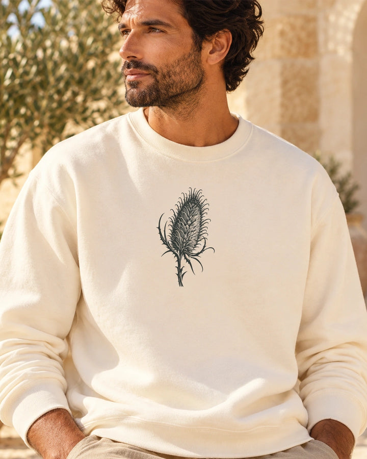 Cardó Ivory lightweight crewneck on a male model in a Mediterranean courtyard, exact locked charcoal teasel artwork centered on front