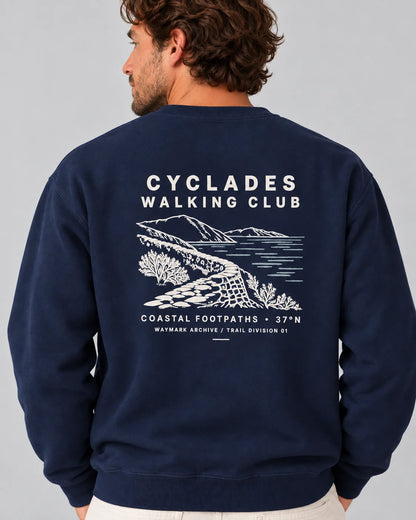 TP HERO: A man wearing a navy blue sweatshirt, viewed from the back. The sweatshirt features a white graphic print of a coastal landscape with the text 'CYCLADES WALKING CLUB' and 'COASTAL FOOTPATHS 37°N'
