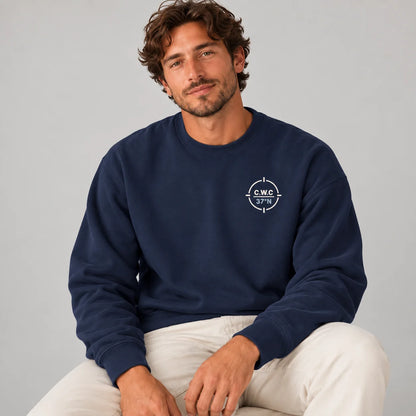 A man wearing a navy blue crewneck sweatshirt with a white compass logo on the left chest, paired with off-white pants, seated against a light gray background.