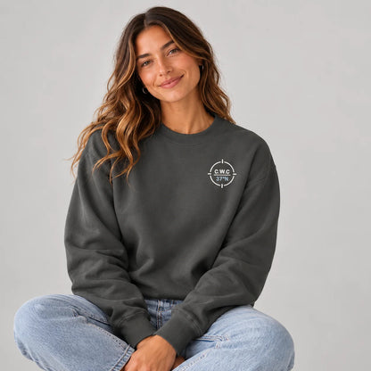 A woman with long wavy brown hair sits cross-legged against a light gray studio background, wearing a dark charcoal crewneck sweatshirt with a small white circular graphic on the left chest and light blue jeans.