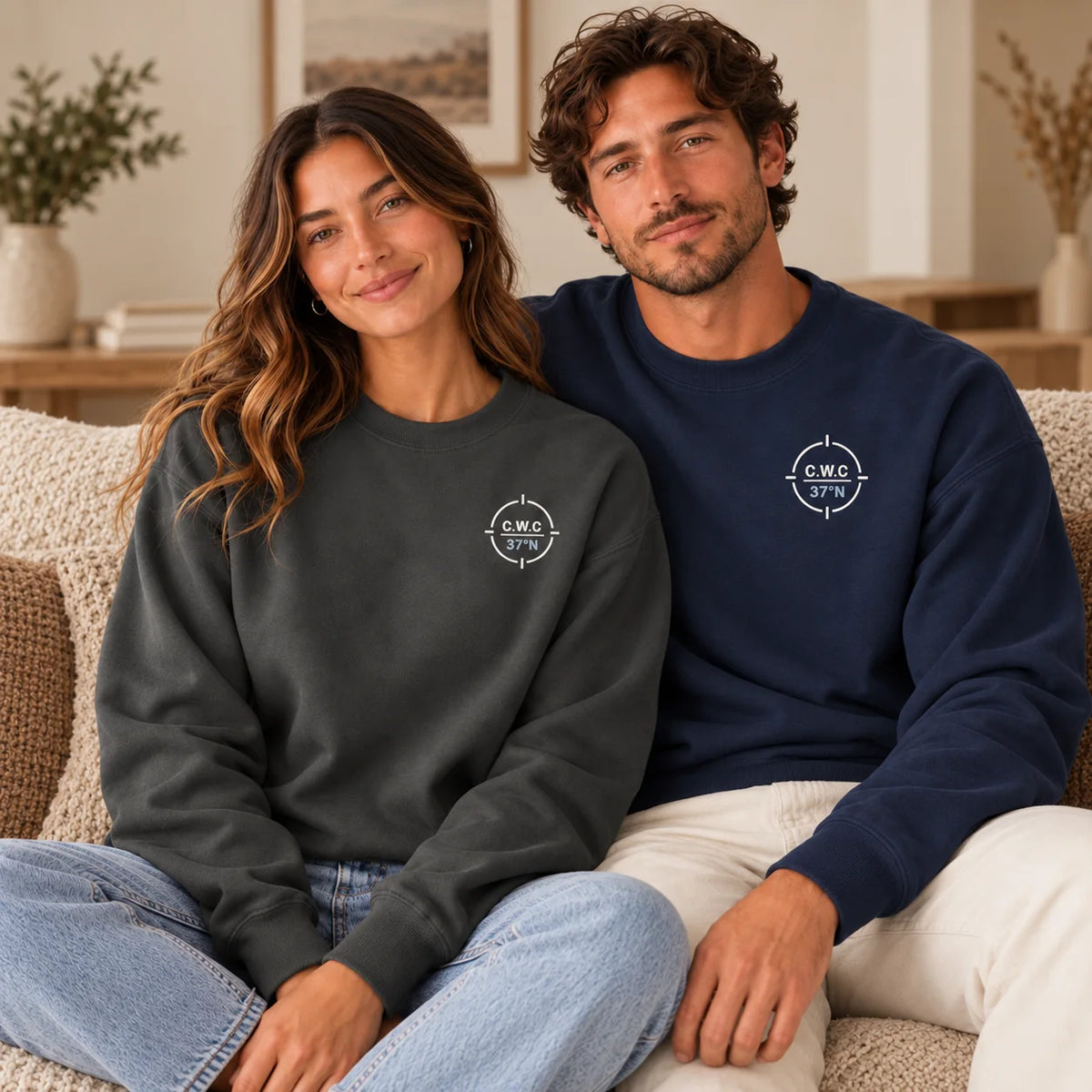 Two models sitting on a beige couch wearing matching crewneck sweatshirts. The model on the left wears a dark charcoal gray sweatshirt, and the model on the right wears a navy blue sweatshirt.