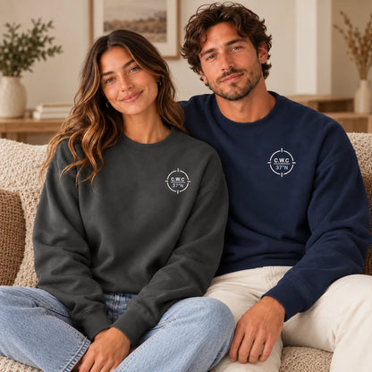 Two models sitting on a beige couch wearing matching crewneck sweatshirts. The model on the left wears a dark charcoal gray sweatshirt, and the model on the right wears a navy blue sweatshirt.