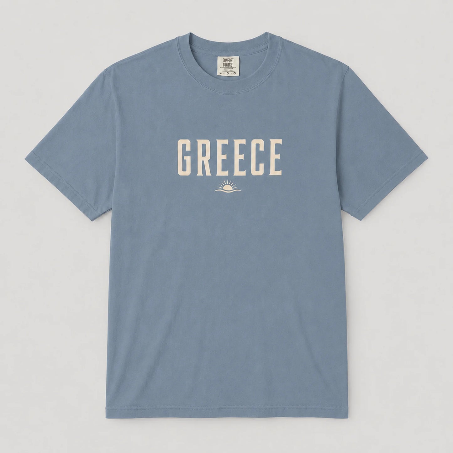 A dusty blue crewneck t-shirt with 'GREECE' printed in cream-colored block letters across the chest, accompanied by a small sun and wave graphic underneath, displayed flat against a light gray background.
