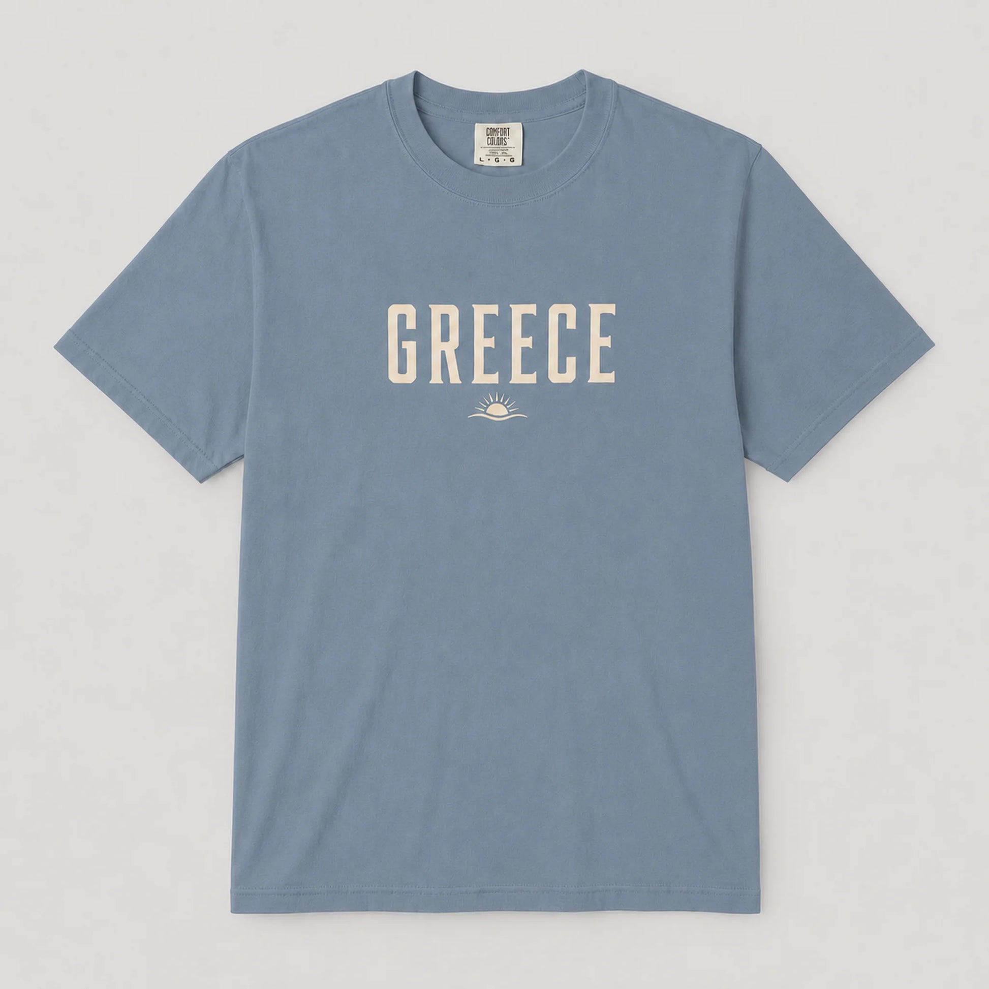 A dusty blue crewneck t-shirt with 'GREECE' printed in cream-colored block letters across the chest, accompanied by a small sun and wave graphic underneath, displayed flat against a light gray background.
