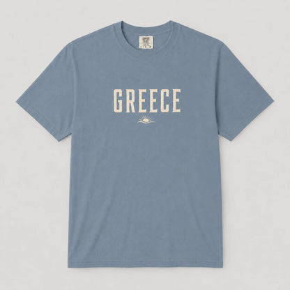 A dusty blue crewneck t-shirt with 'GREECE' printed in cream-colored block letters across the chest, accompanied by a small sun and wave graphic underneath, displayed flat against a light gray background.