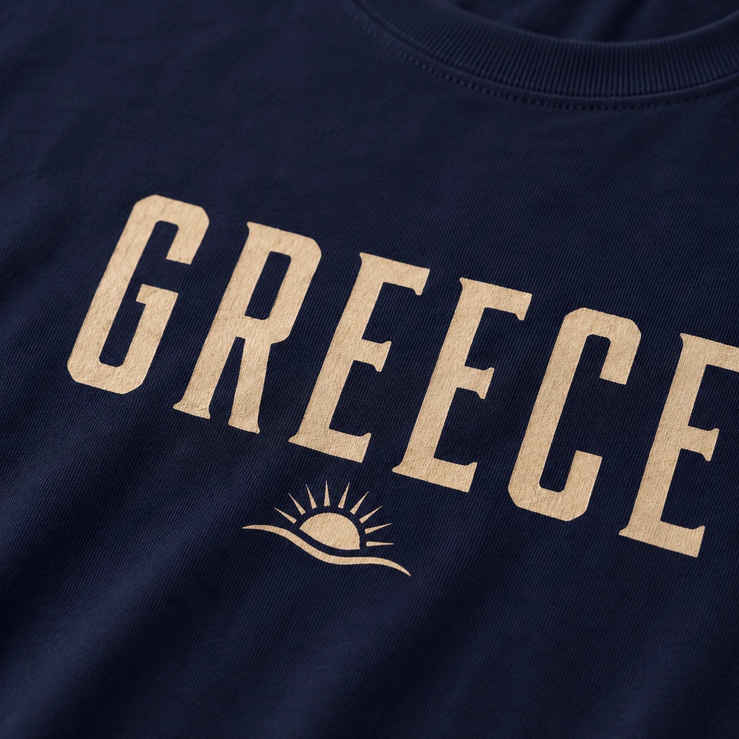 Close-up of a navy blue t-shirt featuring the word 'GREECE' printed in a light beige serif font, with a small sun graphic underneath.