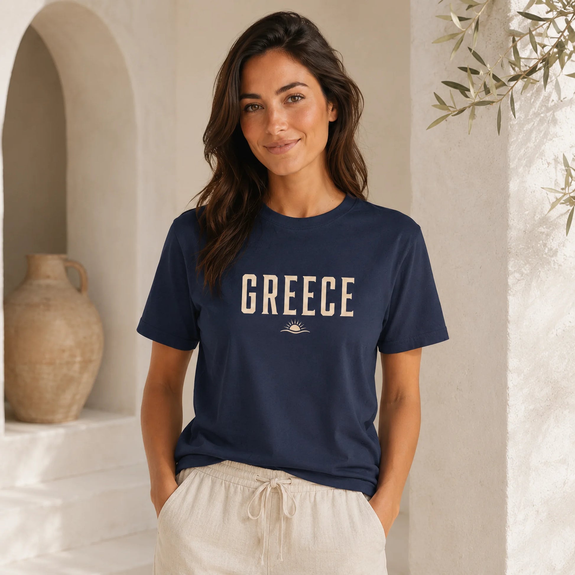 A woman wearing a navy blue t-shirt with 'GREECE' printed in beige across the chest, paired with light beige drawstring pants, standing in a sunlit, rustic interior with a terracotta vase and archway.