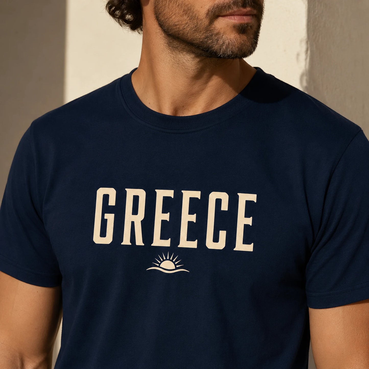 TP HERO: A man wearing a navy blue crewneck t-shirt with 'GREECE' printed in cream block letters and a small sun-and-wave graphic below, set against a warm-toned wall background.