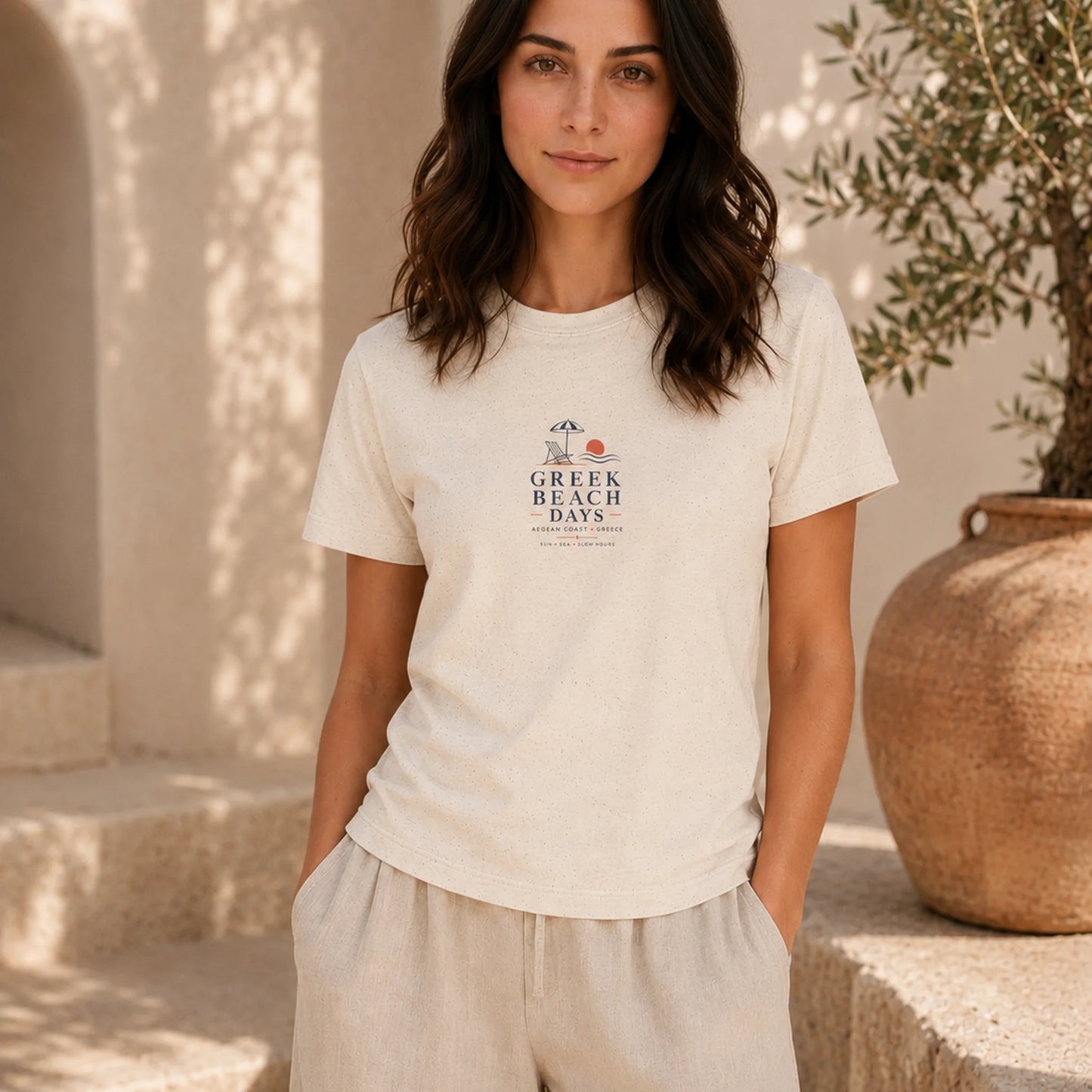 A woman wearing a cream-colored t-shirt with a 'Greek Beach Days' graphic on the chest, paired with matching beige linen pants, standing in a sunlit outdoor setting with stone steps and a large clay pot.