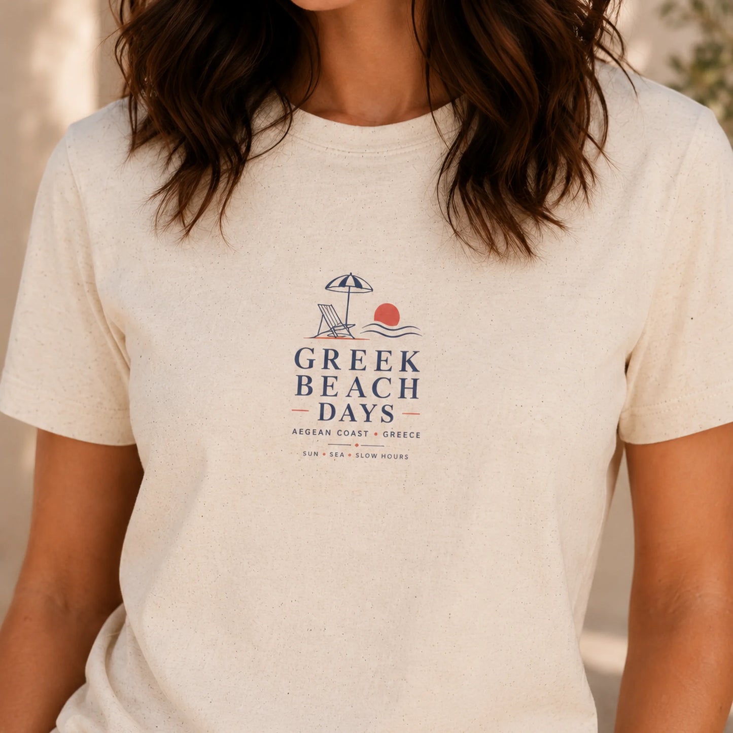 TP HERO: A woman wearing a cream-colored short-sleeve t-shirt with a 'Greek Beach Days' graphic printed on the chest, set against a softly blurred outdoor background.