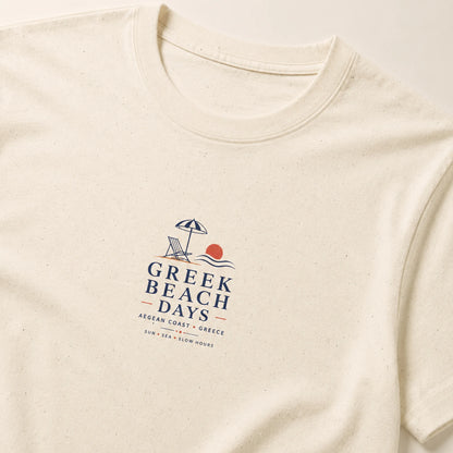 An off-white short-sleeve t-shirt with a textured speckled finish, featuring a 'Greek Beach Days' graphic on the chest with a beach umbrella, chair, waves, and sun.