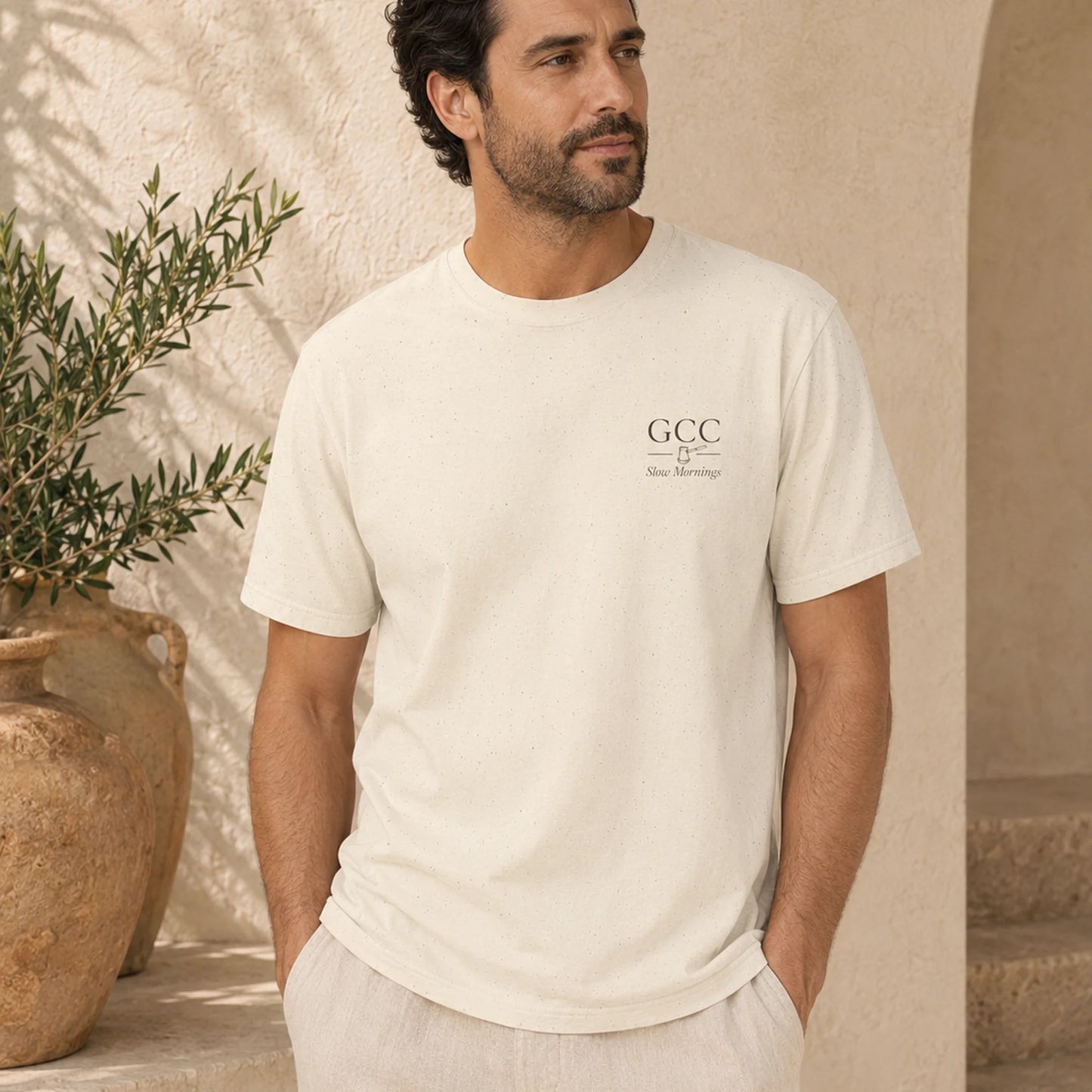 A man wearing a cream-colored short-sleeve t-shirt with a small black 'GCC' logo on the left chest, standing in a warm, sunlit room with beige walls and a potted olive tree.